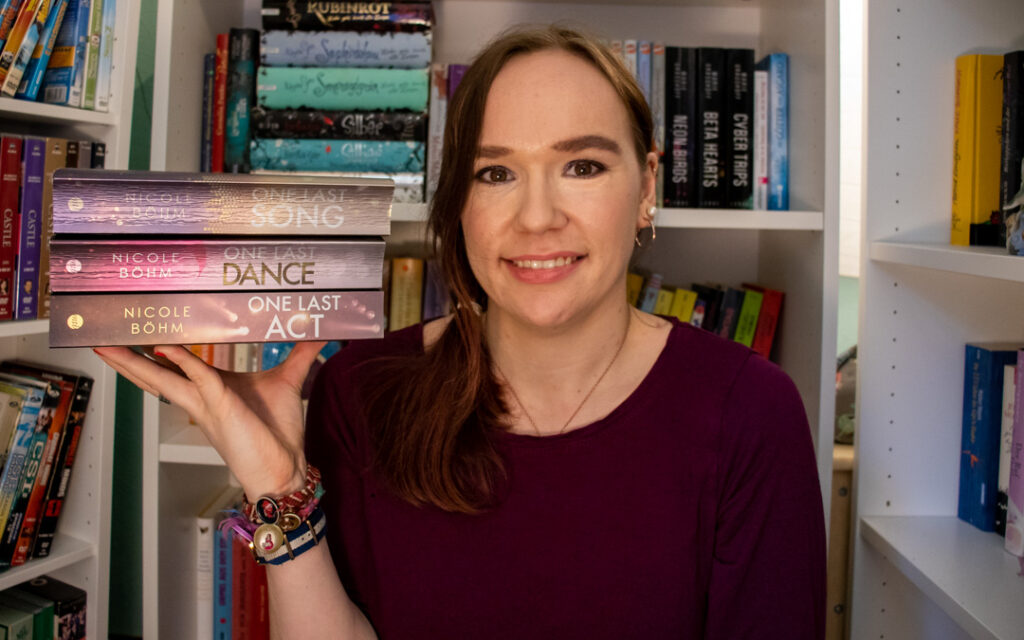 Sue sitzt vor Bücherregalen und hält die drei Bücher der Reihe aufeinander gestapelt auf ihrer Hand mit dem Buchrücken in die Kamera