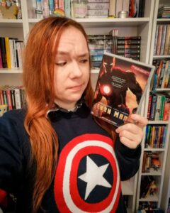 Sue steht vor ihrem Bücherregal in einem Captain America-Pullover und guckt mit fragendem Gesicht auf eine Iron Man-DVD, die sie in der linken Hand hält