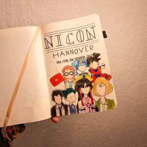 Auf dem Bild sieht man ein Notizbuch, das von links ins Bild gehalten wird. Auf der aufgeschlagenen Seite steht "NiCon Hannover". Darunter steht "07./08. Oktober 2023". In der unteren Hälfte sind 7 Anime-Figuren und das rot-weiße YouTube-Logo zu sehen.