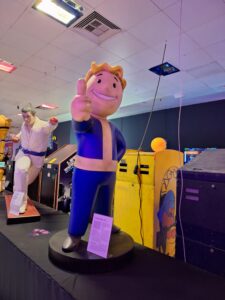 Ein PipBoy aus "Fallout 4" streckt eine Hand mit erhobenem Daumen nach oben. Im Hintergrund sieht man weitere Elemente des Museums.