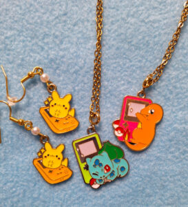 Auf hellblauem Hintergrund sieht man links zwei Ohrhänger mit einem orangen Gameboy und einem Pikachu. In der Mitte ist ein grüner Gameboy mit einem Bisasam und einem kleinen Pokeball. Rechts ist ein pinker Gameboy mit einem Glumanda und zwei Pokebällen.