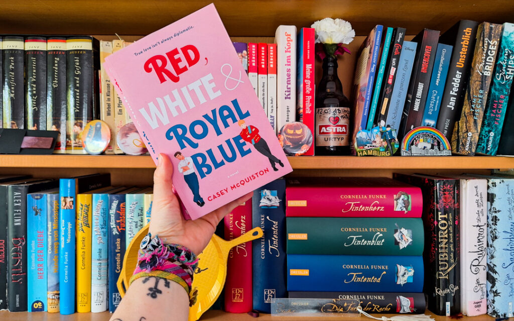 Sue hält das Buch vor ihr Bücherregal mit der linken Hand. Man sieht auf Sue's Arm ein Anker-Tattoo und viele bunte selbstgeknüpfte Armbänder am Handgelenk. Das Buch hält sie leicht schräg nach links geneigt. Auf dem rosanen Cover sieht man den Titelschriftzug in vier Zeilen. Die Wörter von "Red, White & Royal Blue" sind jeweils eingefärbt in die benannte Farbe. Links neben dem Blue von Royal Blue steht Alex in Jeans und weißem Hemd, rechts davon steht Henry in einer Uniform des britischen Königshauses.